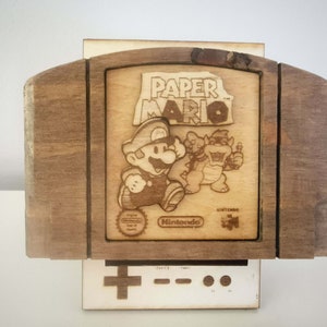 Könnte beinhalten: Hölzerne Nachbildung der Paper Mario Nintendo 64-Spielkassette. Der Spieletitel und die Charaktere Mario und Bowser sind in das Holz geätzt. Die Kassette sitzt auf einer weißen Controllerbasis mit einem kreuzförmigen Steuerkreuz.