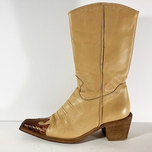 Könnte beinhalten: Beigefarbene Cowboy-Stiefel aus Leder mit spitzer Zehenkappe und braunem Ledereinsatz an der Spitze. Die Stiefel haben einen Blockabsatz.