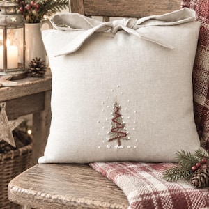 Könnte beinhalten: Ein quadratisches beiges Leinenkissen mit einer dekorativen Stoffschleife an der Oberseite und einem gestickten Weihnachtsbaummotiv in der Mitte. Der Baum ist mit grünem und rotem Garn gestickt und von kleinen weißen Knotenpunkten umgeben.