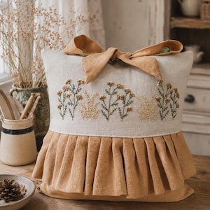 May include: A square throw pillow featuring a light beige linen front with embroidered yellow and green wildflower motifs. The pillow is accented with a mustard-colored fabric bow at the top and a matching mustard-colored pleated ruffle trim.