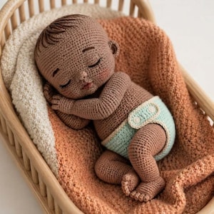 Könnte beinhalten: Eine handgefertigte Häkelpuppe eines schlafenden Säuglings mit dunkelbrauner Haut, texturiertem Haar und rosigen Wangen. Die Puppe trägt eine hellminzgrüne Windel mit zwei weißen Knöpfen an der Seite und liegt in einer kleinen hölzernen Wiege.
