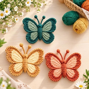 May include: Three crocheted butterfly appliques in teal, yellow, and orange color schemes. Each butterfly features a central body, two antennae, and detailed wing patterns with contrasting inner and outer yarn colors, crafted with visible stitch texture.