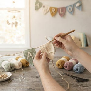 May include: A person crocheting a triangular pennant with the letter L in cream yarn, attached to a string with a sage green pennant featuring the letter A. Several balls of yarn in sage, cream, mustard, pink, and blue sit nearby with scissors.