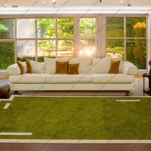 May include: A modern living room featuring a white sofa with mustard-colored pillows, a green rug with a white border, and large windows overlooking a lush outdoor scene. The rug has a unique geometric design.