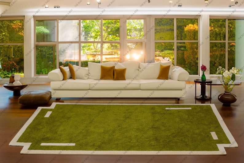 May include: A modern living room featuring a white sofa with mustard-colored pillows, a green rug with a white border, and large windows overlooking a lush outdoor scene. The rug has a unique geometric design.
