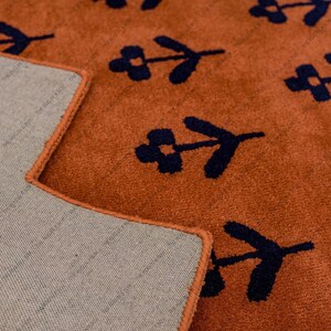 May include: Close-up of a terracotta-colored rug with a repeating pattern of navy blue floral motifs. The rug's edge is neatly finished with a matching terracotta border. The underside of the rug is a neutral beige.