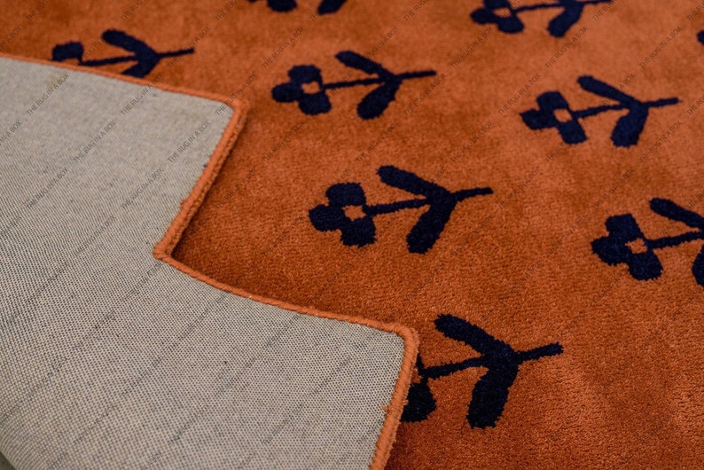 May include: Close-up of a terracotta-colored rug with a repeating pattern of navy blue floral motifs. The rug's edge is neatly finished with a matching terracotta border. The underside of the rug is a neutral beige.