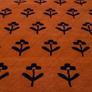 May include: Close-up of a burnt orange rug with a repeating pattern of navy blue stylized flowers. The rug has a soft, textured appearance, and the floral design is evenly spaced across the surface. The rug is a decorative home decor item.