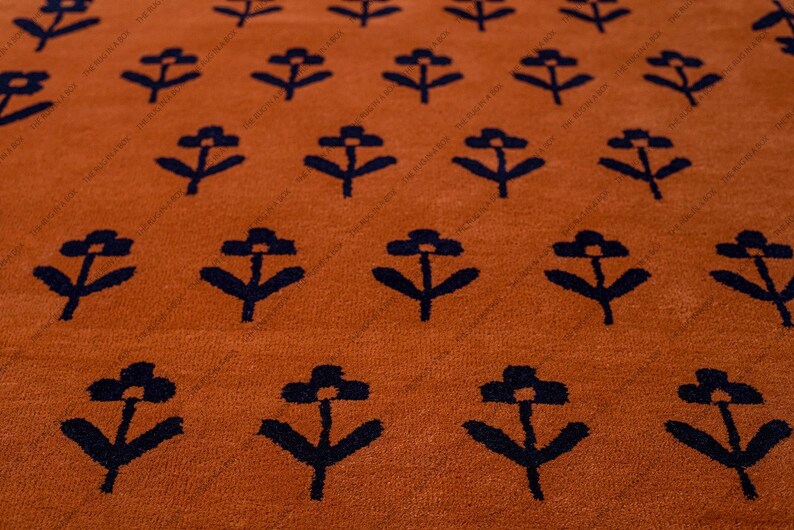 May include: Close-up of a burnt orange rug with a repeating pattern of navy blue stylized flowers. The rug has a soft, textured appearance, and the floral design is evenly spaced across the surface. The rug is a decorative home decor item.
