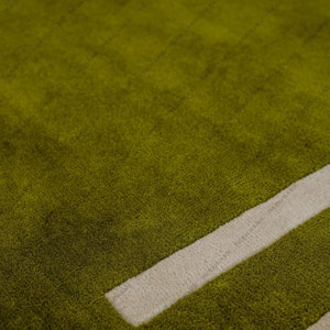 May include: Close-up of a green rug with a soft, textured surface. A thick, off-white rectangular design element is visible, adding a contrasting geometric detail to the rug's overall design. The rug's color is a vibrant, earthy green.