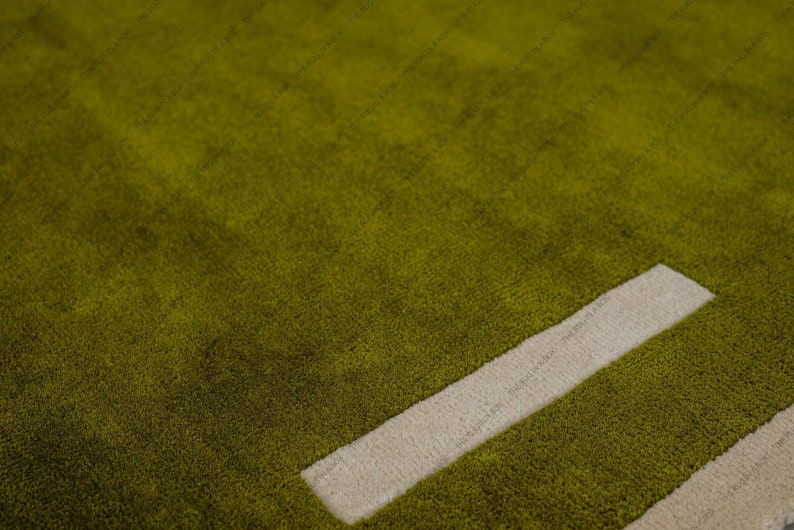 May include: Close-up of a green rug with a soft, textured surface. A thick, off-white rectangular design element is visible, adding a contrasting geometric detail to the rug's overall design. The rug's color is a vibrant, earthy green.