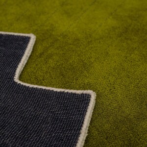 May include: Close-up of a green rug with a dark blue backing. The rug has a textured surface and a cream-colored border. The backing is visible in the lower left corner, showing the woven texture.