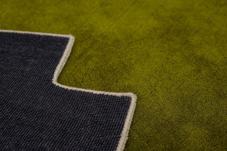 May include: Close-up of a green rug with a dark blue backing. The rug has a textured surface and a cream-colored border. The backing is visible in the lower left corner, showing the woven texture.