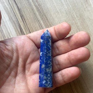Puede incluir: Una punta de cristal de lapislázuli pulido con forma hexagonal. La piedra presenta un color base azul real profundo con motas visibles de pirita dorada e inclusiones de calcita blanca por toda la superficie de la torre facetada.