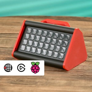 Könnte beinhalten: Ein rot-schwarzes elektronisches Gerät mit einem Raster aus Knöpfen. Das Gerät hat einen Griff oben und eine dreieckige Form an der Seite.