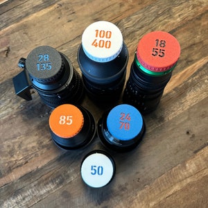 Op de afbeelding: Zes verschillende lensdoppen met bedrukte nummers. De lensdoppen zijn wit, oranje, blauw en rood. De nummers op de lensdoppen zijn 100 400, 28 135, 85, 24 70, 50 en 18 55.