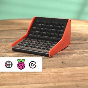 May include: A black and orange electronic device with a grid of small, square buttons. The device has a sloped design with orange side panels and a black top. The device is on a wooden surface.