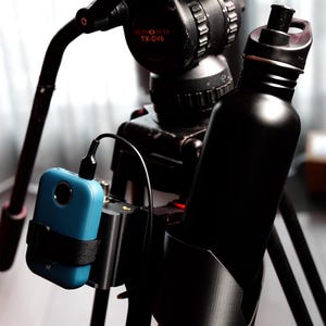 May include: A black tripod with a camera mounted on top. A blue device is attached to the side of the tripod, and a black water bottle is attached to the other side. The camera has a red label that says "TX-DV6".