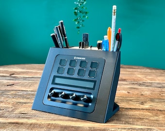 Può includere: Un Stream Deck mini nero con otto pulsanti, un piccolo schermo e quattro manopole. Il dispositivo è su un tavolo di legno e ha un portapenne con penne e matite.