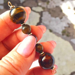 May include: A string of polished gemstone beads in shades of brown and gold. The beads vary in size and are strung together with silver-coloured metal accents and gold-coloured loops. The largest bead has a tiger's eye pattern.