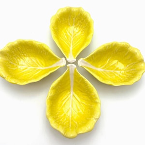 May include: Four yellow ceramic leaf-shaped dishes arranged in a flower-like pattern. Each dish has a central white vein design, with a bright yellow exterior. The dishes are likely for serving appetizers or small portions.