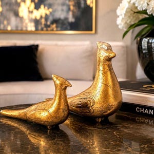 May include: Two gold-colored bird sculptures with textured surfaces. One bird is lying down, and the other is upright. The sculptures are on a dark, patterned surface, with a couch and artwork in the background. Home decor.