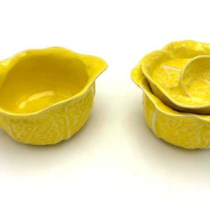 May include: A bright yellow ceramic creamer and covered sugar bowl set. The creamer has a handle and a spout. The sugar bowl is shaped like a cabbage with a lid. Both pieces have a textured surface and are on a white background.