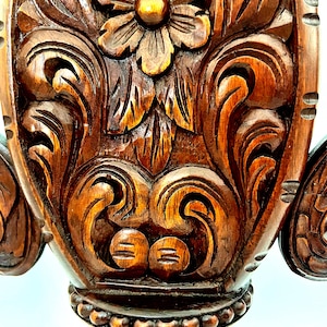 Puede incluir: Primer plano de un detalle de mueble de madera ricamente tallado. La madera marr&oacute;n oscuro presenta intrincados dise&ntilde;os florales y de volutas, con una flor prominente en el centro. El tallado tiene un efecto tridimensional, mostrando la artesan&iacute;a.