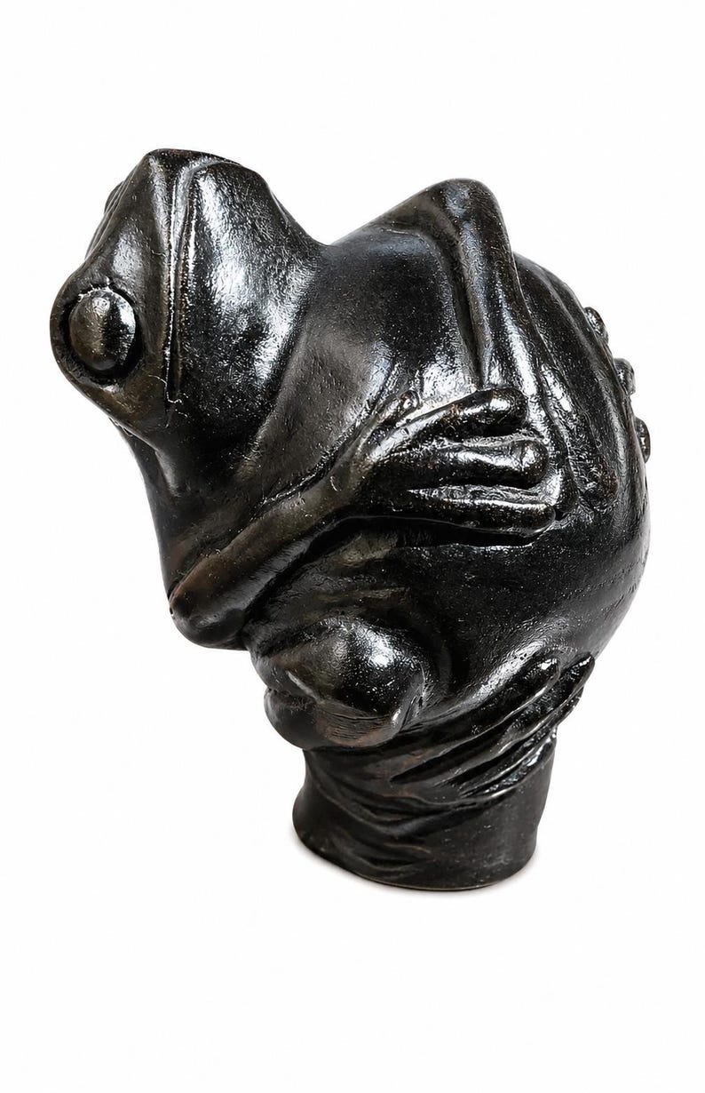 May include: A dark bronze frog sculpture with a textured surface. The frog is in a seated position with its arms crossed. The sculpture has a detailed design and is set against a white background.