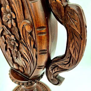 Puede incluir: Primer plano de un jarr&oacute;n de madera tallada de color marr&oacute;n oscuro. El jarr&oacute;n presenta dise&ntilde;os florales y de hojas, con un asa decorativa. La base es redonda y ornamentada, con un patr&oacute;n detallado. La madera tiene un tono rico y c&aacute;lido.