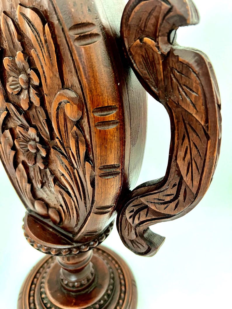 Puede incluir: Primer plano de un jarr&oacute;n de madera tallada de color marr&oacute;n oscuro. El jarr&oacute;n presenta dise&ntilde;os florales y de hojas, con un asa decorativa. La base es redonda y ornamentada, con un patr&oacute;n detallado. La madera tiene un tono rico y c&aacute;lido.