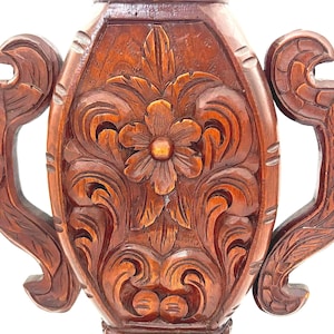 Puede incluir: Primer plano de un jarr&oacute;n de madera tallada con un dise&ntilde;o floral. El jarr&oacute;n es de color marr&oacute;n intenso y presenta intrincadas tallas de flores y hojas. El jarr&oacute;n tiene dos asas a cada lado.