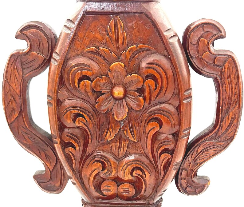 Puede incluir: Primer plano de un jarr&oacute;n de madera tallada con un dise&ntilde;o floral. El jarr&oacute;n es de color marr&oacute;n intenso y presenta intrincadas tallas de flores y hojas. El jarr&oacute;n tiene dos asas a cada lado.