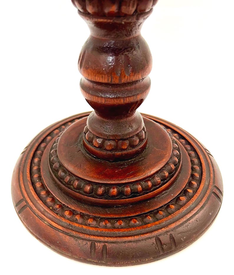 Puede incluir: Primer plano de un pedestal de madera tallada de color marr&oacute;n oscuro. El pedestal tiene una base redonda con un borde de cuentas y una columna torneada detallada. La madera tiene un acabado rico y pulido, mostrando las intrincadas tallas.