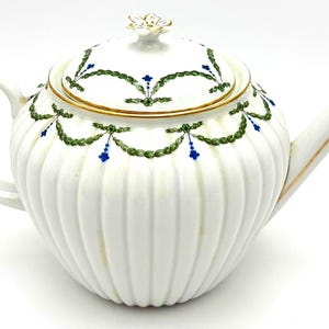 May include: A white porcelain teapot with a ribbed body, featuring a delicate green garland pattern and small blue floral accents. The lid is topped with a white flower-shaped finial, and the edges are finished with a thin gold trim.