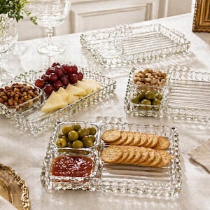 May include: A collection of clear, decorative glass serving trays and dishes. The trays are filled with snacks like grapes, cheese, olives, crackers, and nuts. The trays have a scalloped edge and are arranged on a white tablecloth.