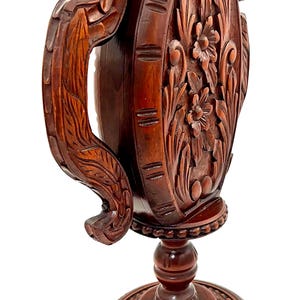 Puede incluir: Un jarr&oacute;n de madera marr&oacute;n oscuro, tallado intrincadamente, con base de pedestal y asa. El jarr&oacute;n presenta dise&ntilde;os florales y de hojas, y el asa tiene una forma curva detallada. La base es redonda y tambi&eacute;n tiene detalles tallados.