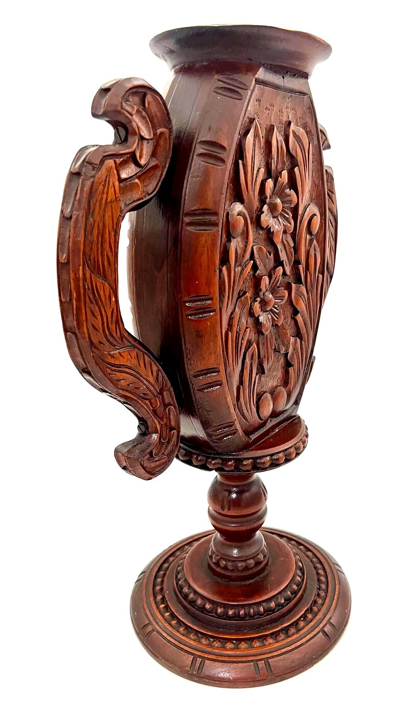 Puede incluir: Un jarr&oacute;n de madera marr&oacute;n oscuro, tallado intrincadamente, con base de pedestal y asa. El jarr&oacute;n presenta dise&ntilde;os florales y de hojas, y el asa tiene una forma curva detallada. La base es redonda y tambi&eacute;n tiene detalles tallados.
