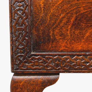 May include: Close-up of a section of a wooden antique table. The table features a dark brown finish with intricate carved knotwork along the edges. The corner of the table leg is visible, showcasing the craftsmanship and age of the furniture.