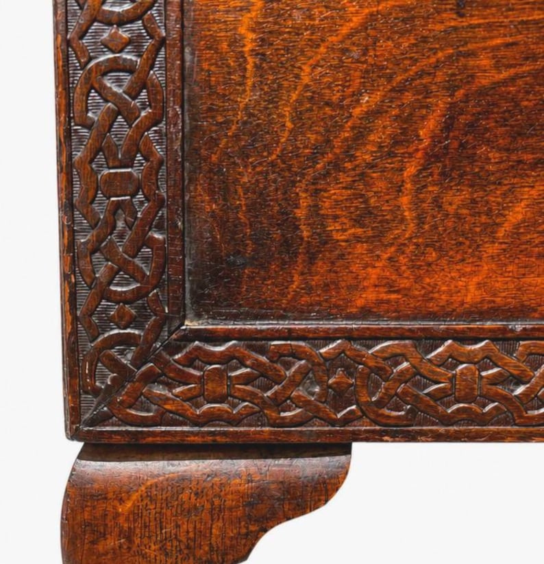 May include: Close-up of a section of a wooden antique table. The table features a dark brown finish with intricate carved knotwork along the edges. The corner of the table leg is visible, showcasing the craftsmanship and age of the furniture.