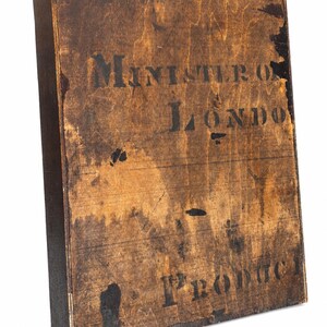 May include: A vintage wooden box with dark brown staining and black stenciled text. The text reads "MINISTERO LONDON" and "1st PRODUCT". The box has a rectangular shape and is likely an antique.