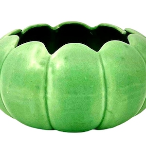 May include: A green ceramic bowl with a pumpkin-like shape. The bowl has a scalloped rim and vertical ridges, giving it a textured appearance. The interior is dark, contrasting with the bright green exterior.