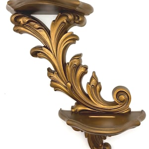 May include: Ornate gold-colored wall shelf with a scrolling leaf design. The shelf has two curved platforms and decorative flourishes. The shelf is made of a solid material and is designed for displaying small items.