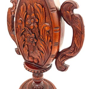 Puede incluir: Un jarr&oacute;n de madera marr&oacute;n oscuro tallado a mano con intrincados dise&ntilde;os florales y de hojas. El jarr&oacute;n presenta dos asas ornamentadas y una base detallada. La madera tiene un acabado pulido, mostrando la veta y textura natural.