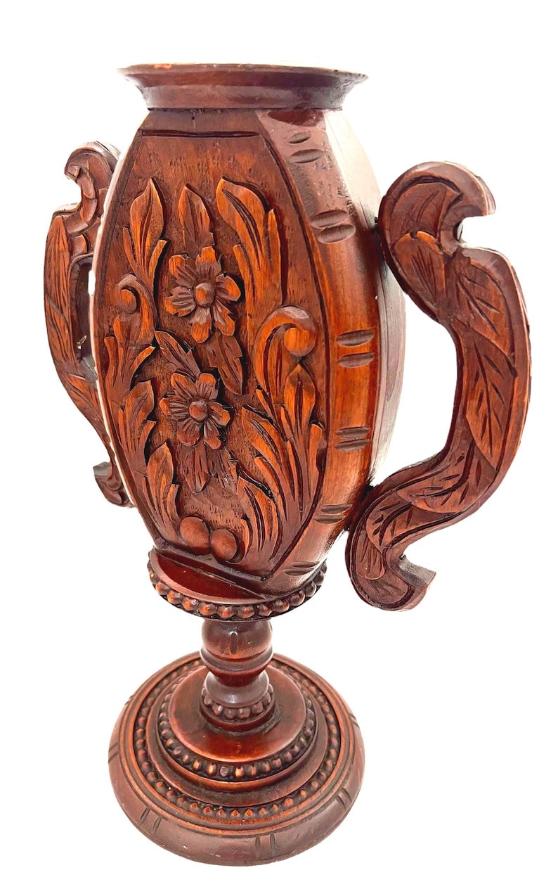 Puede incluir: Un jarr&oacute;n de madera marr&oacute;n oscuro tallado a mano con intrincados dise&ntilde;os florales y de hojas. El jarr&oacute;n presenta dos asas ornamentadas y una base detallada. La madera tiene un acabado pulido, mostrando la veta y textura natural.