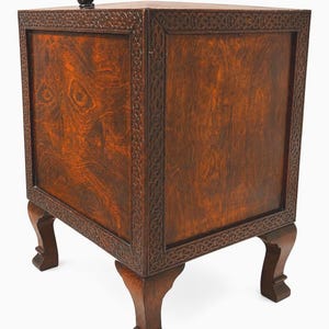 May include: A dark brown wooden side table with carved details and a small, dark sculpture on top. The table has a square shape with four legs and decorative trim around the edges. The wood grain is visible.