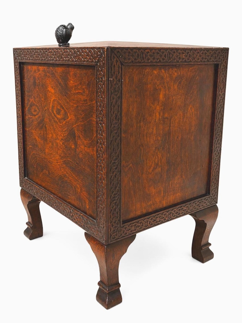 May include: A dark brown wooden side table with carved details and a small, dark sculpture on top. The table has a square shape with four legs and decorative trim around the edges. The wood grain is visible.