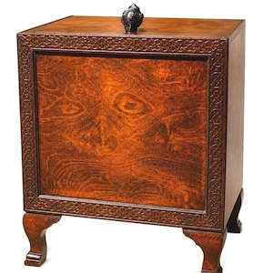 May include: A wooden cabinet with a dark brown finish, featuring a decorative carved border and ornate feet. The cabinet has a small, dark, decorative knob on top. The wood grain is visible on the front panel.