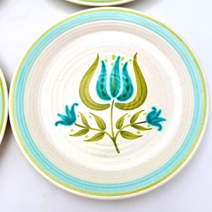 May include: White ceramic plates with a turquoise and green floral design. The plates have a speckled off-white center with a turquoise and green border. The floral design features a large turquoise flower with green leaves and smaller turquoise flowers.