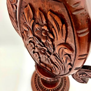 Puede incluir: Primer plano de un objeto decorativo de madera tallada de color marr&oacute;n oscuro. El objeto presenta intrincados dise&ntilde;os florales y de hojas. La base es redonda con un patr&oacute;n detallado. La madera tiene un tono rico y c&aacute;lido, y la talla es muy detallada.
